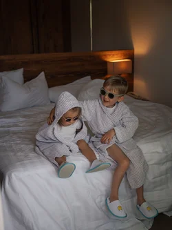 Due bambini con accappatoi e occhiali da sole seduti su un letto in una stanza accogliente