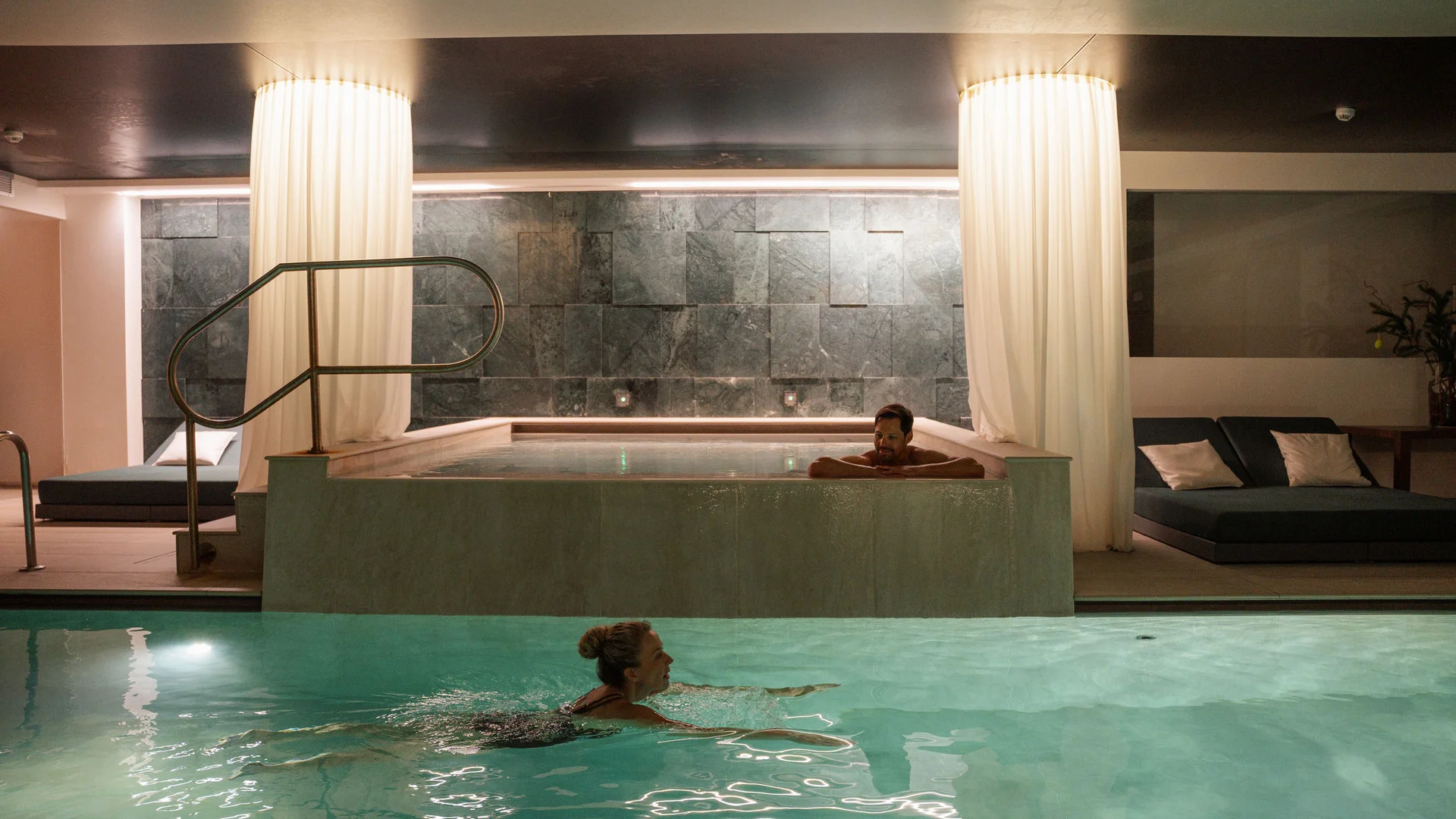 Elgus: hotel a 4 stelle in Trentino Donna che nuota in piscina interna con uomo che si rilassa in vasca idromassaggio