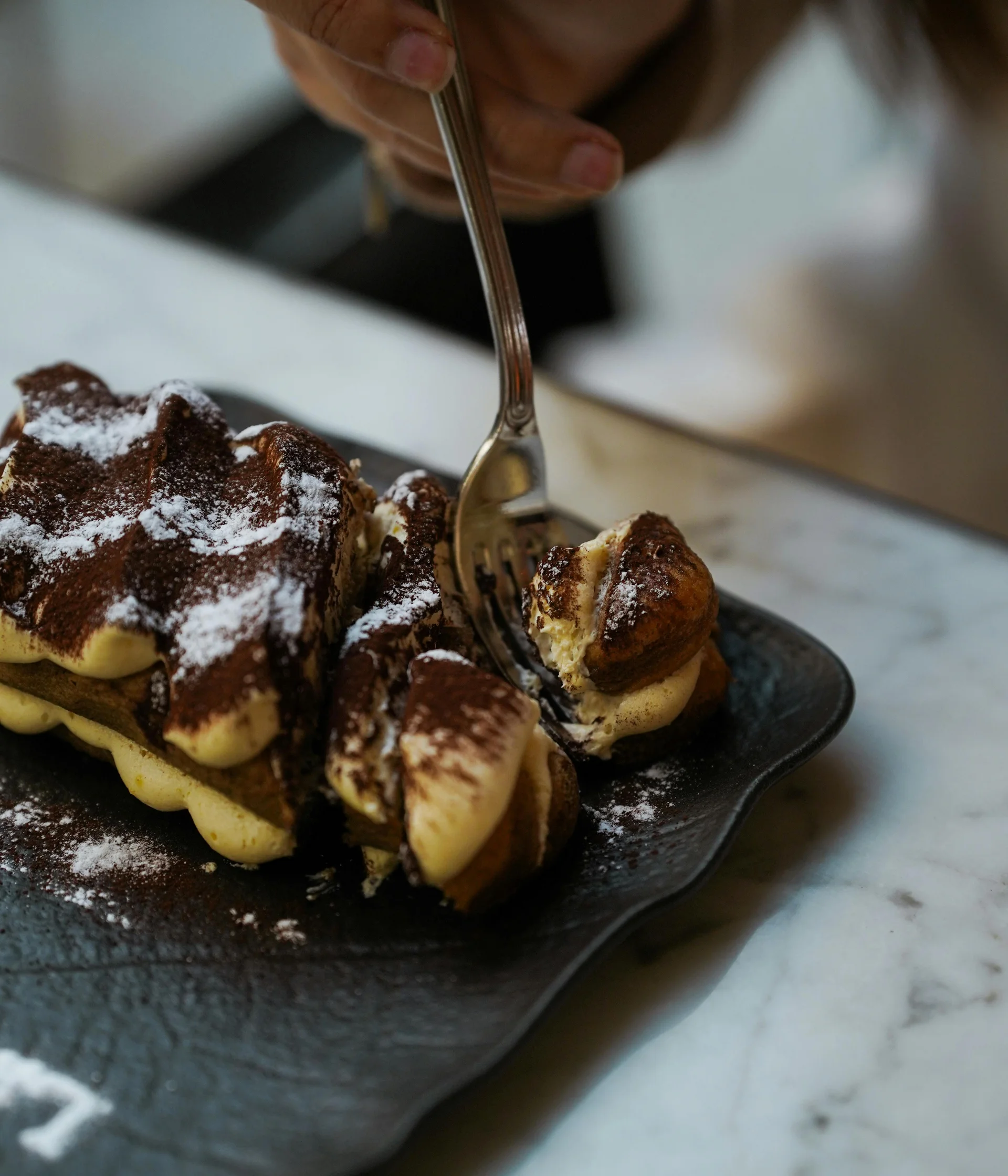 Il nostro ristorante a San Giacomo, Brentonico Mano che prende una fetta di tiramisù con una forchetta su un piatto nero