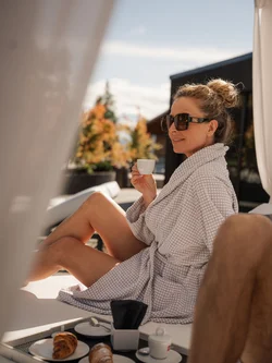 Donna con accappatoio e occhiali da sole che beve caffè all'aperto con croissant