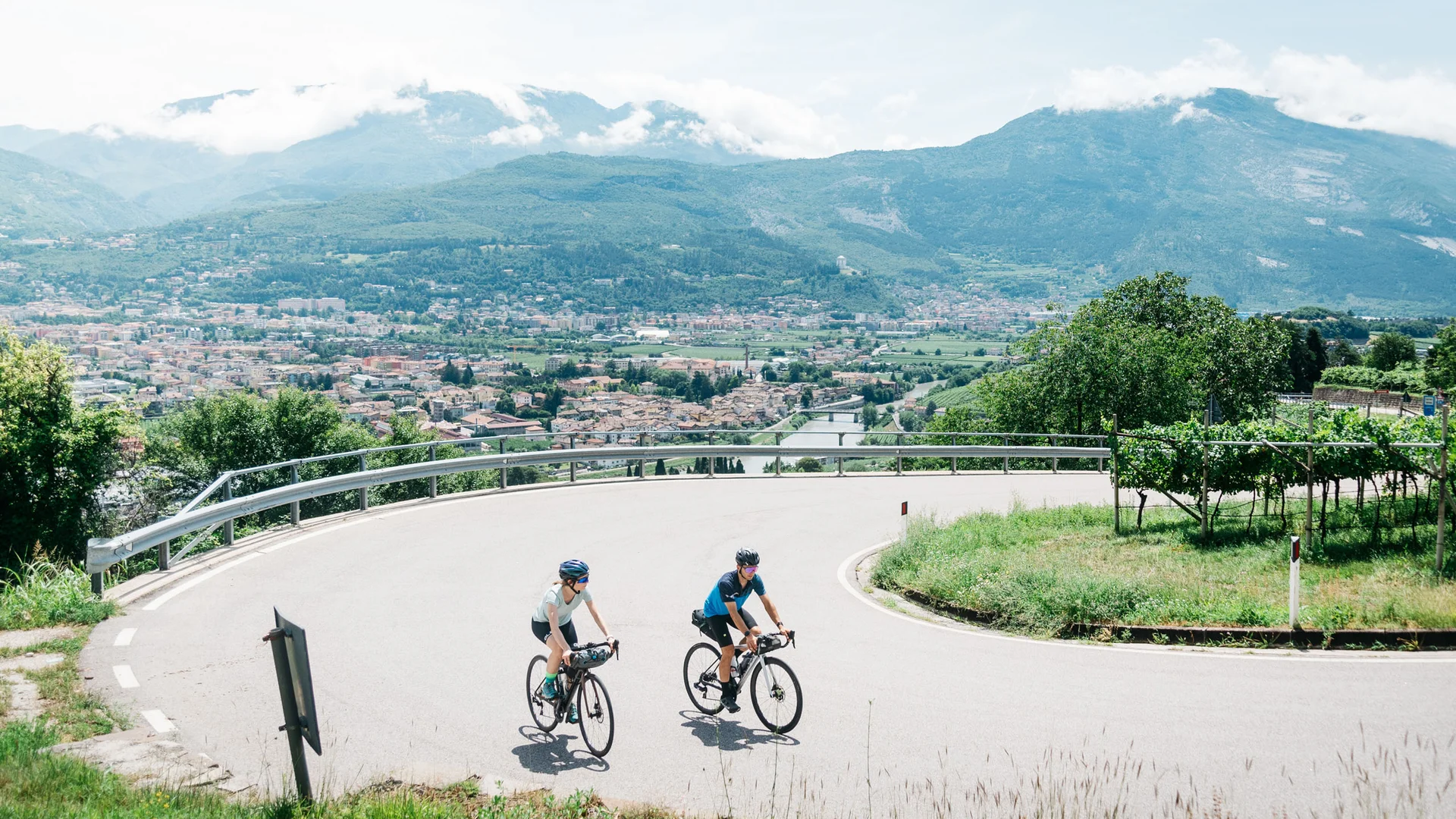 Elgus: hotel a 4 stelle in Trentino Due ciclisti su strada curva con vista panoramica su città e montagne