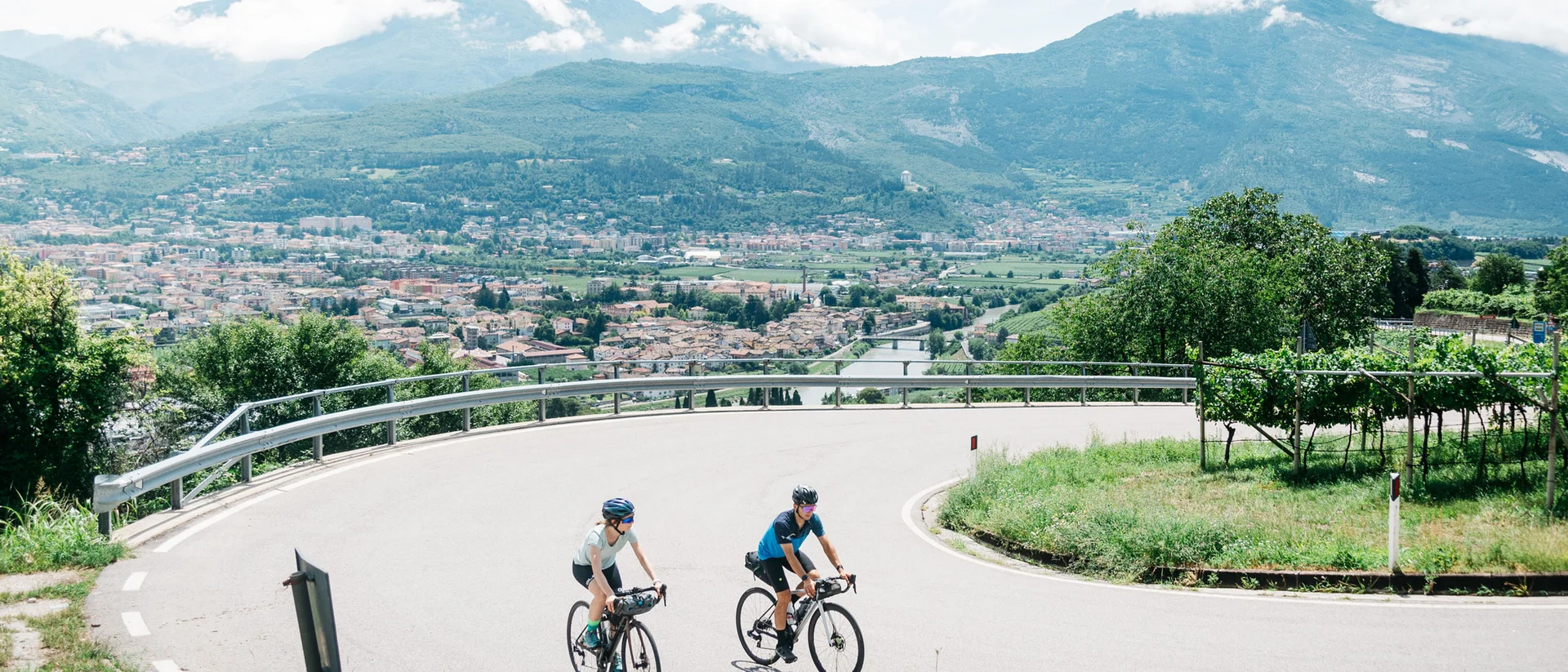 Tutto il divertimento di un hotel sul Monte Baldo Due ciclisti su strada curva con vista panoramica su città e montagne
