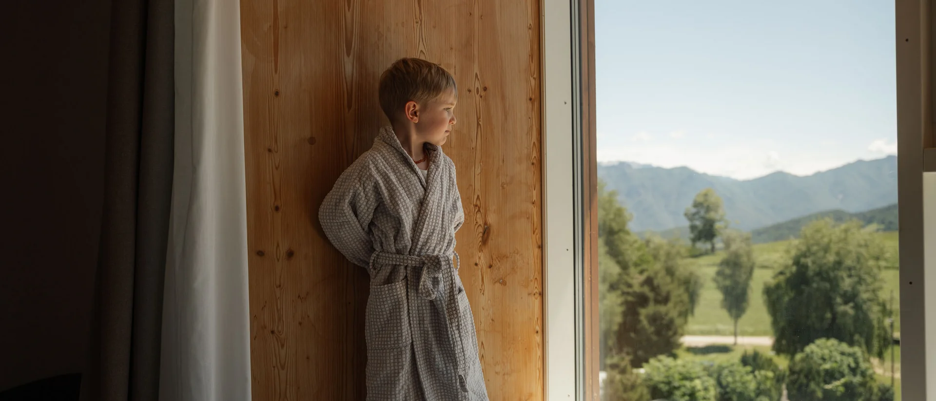 Elgus: family hotel a Brentonico Bambino in accappatoio che guarda fuori dalla finestra in una stanza di legno