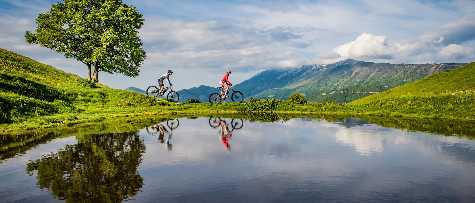 Tutto il divertimento di un hotel sul Monte Baldo Due ciclisti su sentiero verde vicino a lago con riflesso di albero e montagne