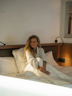 Lächende Frau sitzt im Bett mit Bademantel und weißen Decken