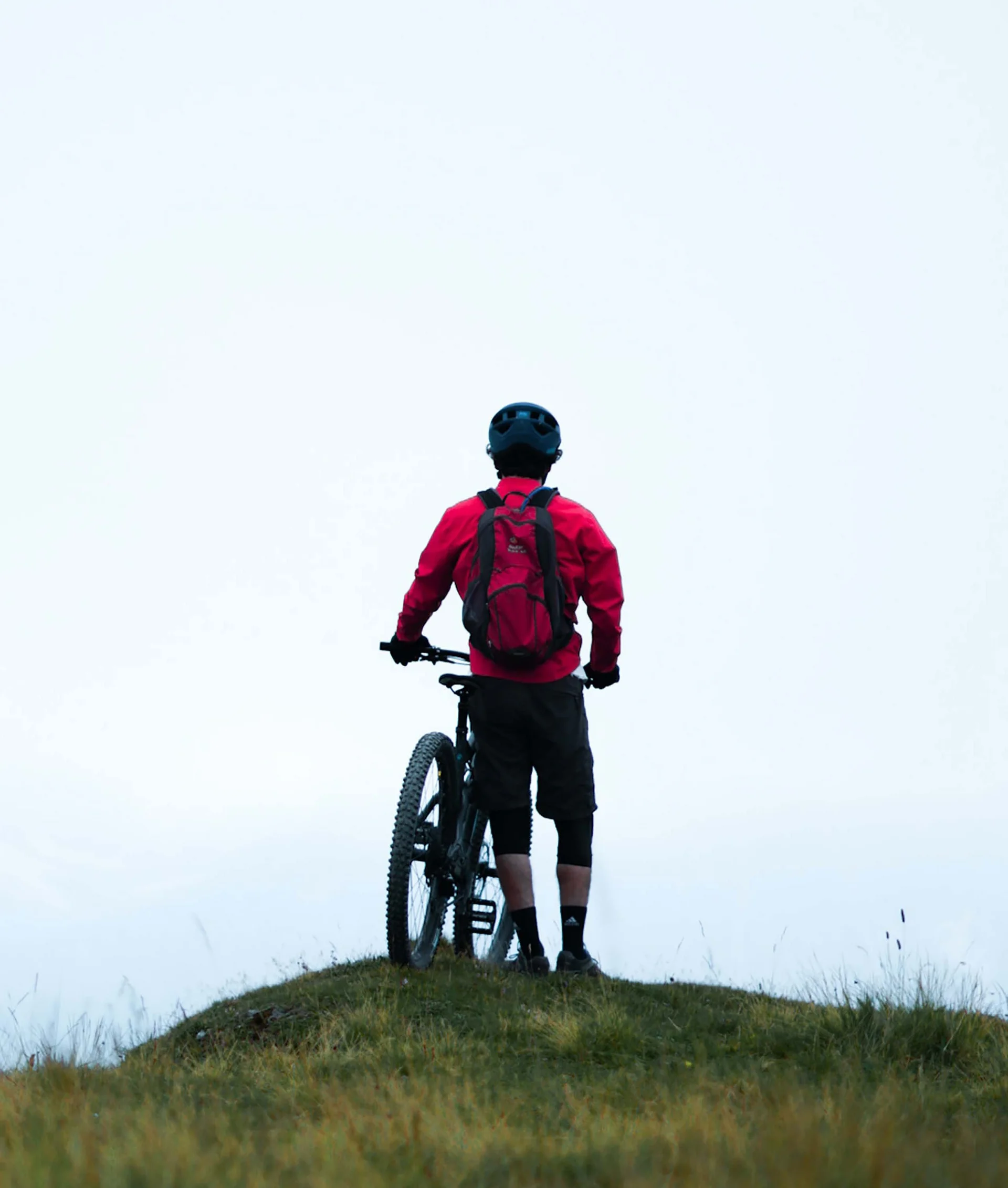 Hotel per single in Trentino Ciclista con giacca rossa e casco in cima a una collina erbosa con bicicletta