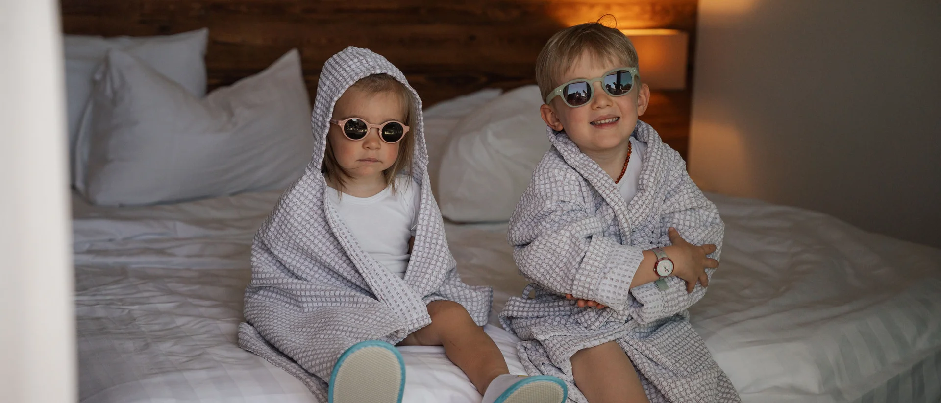 Elgus: hotel con spa per famiglie in Trentino Due bambini con accappatoi e occhiali da sole seduti su un letto in una camera d'hotel